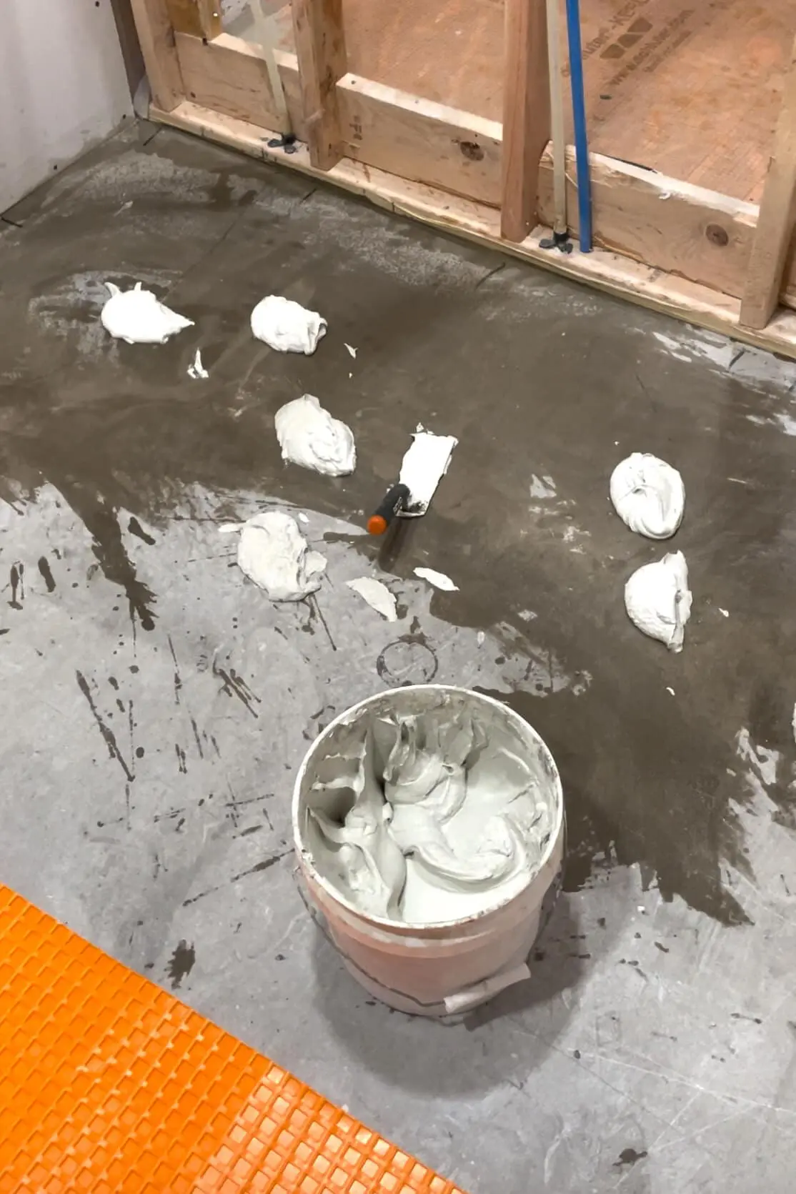 A bathroom floor prepped with dollops of thinset mortar spread across the surface, alongside a trowel and a bucket filled with mixed mortar, ready for underlayment or tile installation.